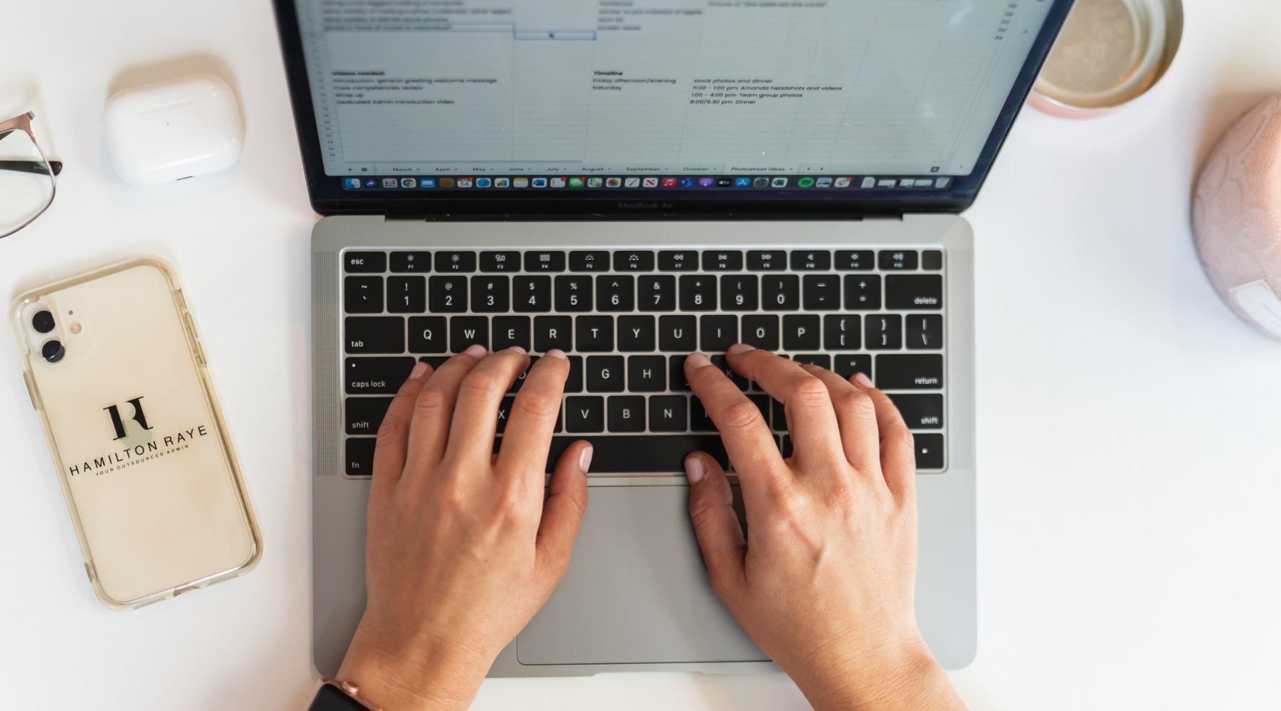 Hands typing on a MacBook keyboard with a Hamilton Raye-branded iPhone case, AirPods, glasses, and a pink mug placed nearby on a white desk.