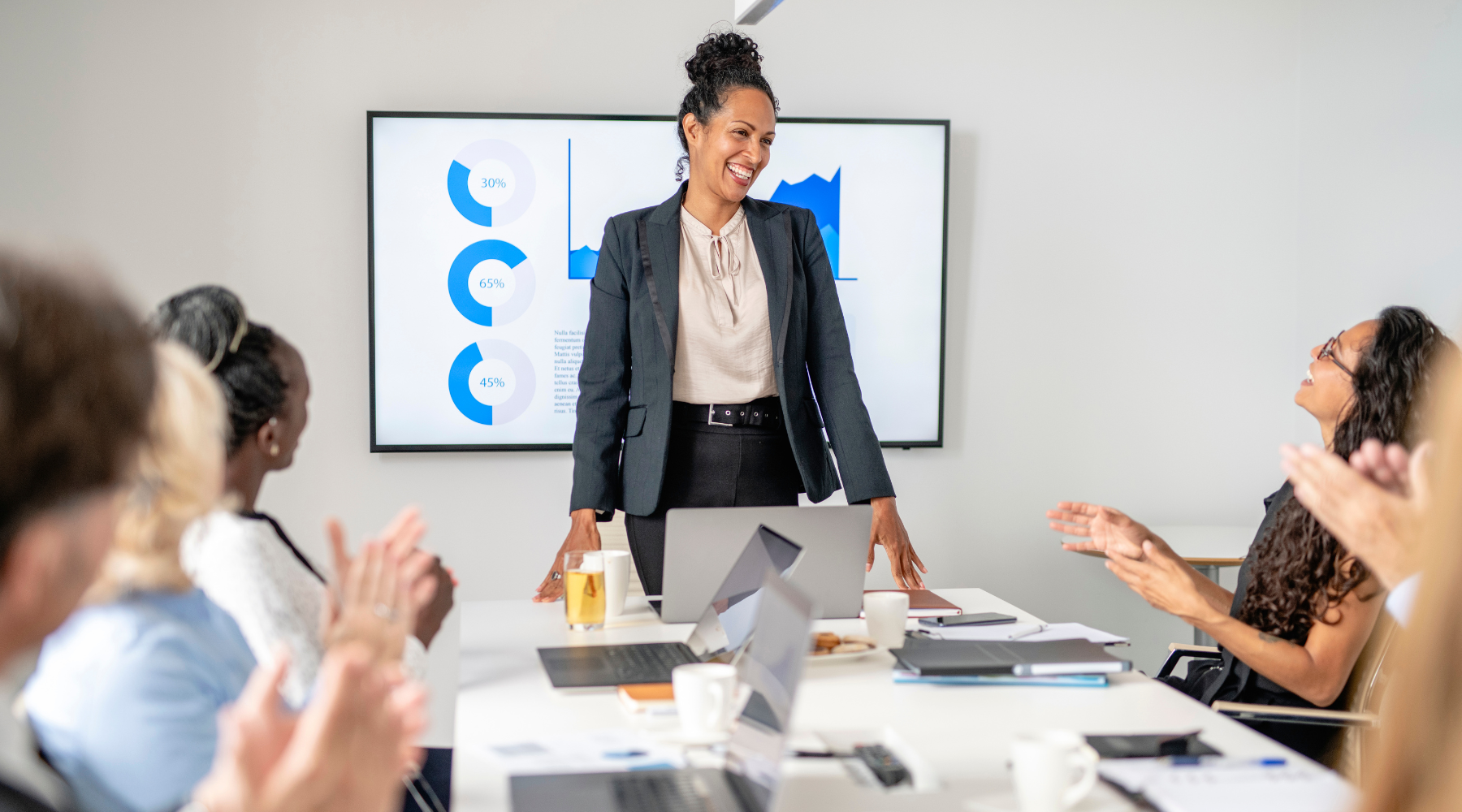 Confident businesswoman leading a meeting, standing at the head of a conference table while team members applaud—illustrating the impact of a strategic partner in leadership.