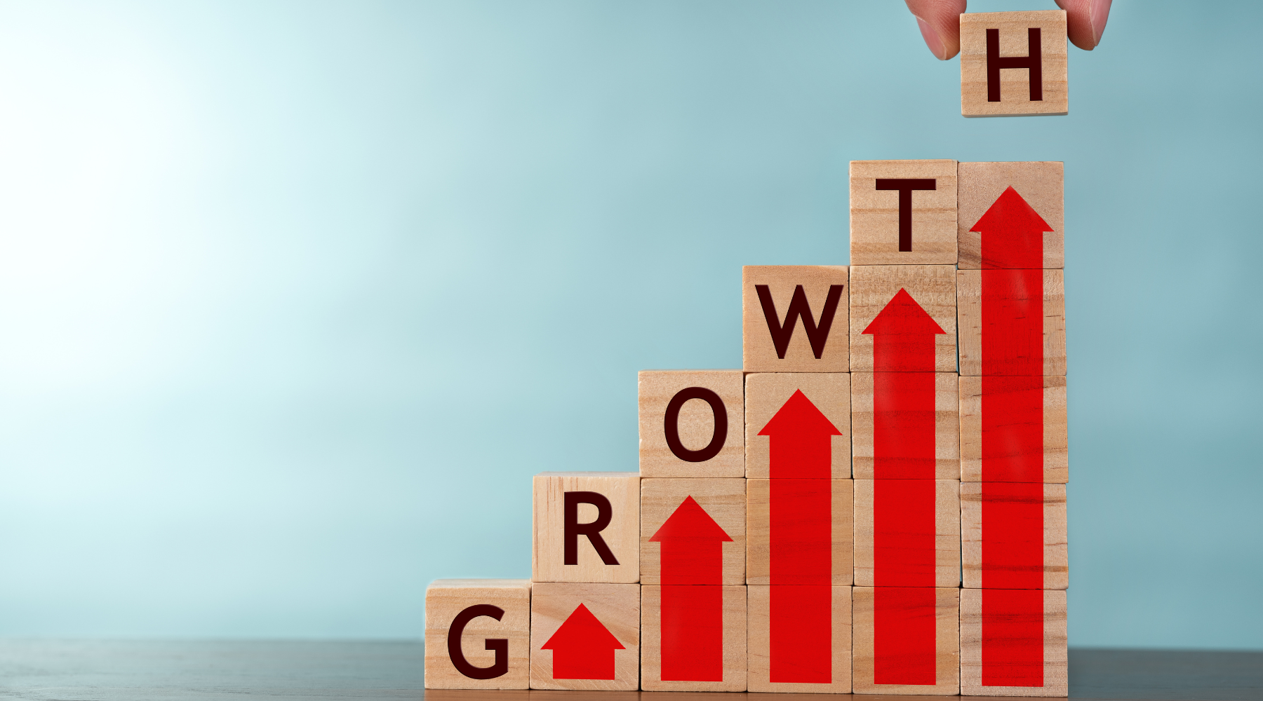 A hand places the final wooden block labeled "H" atop a staircase of blocks spelling out "GROWTH," each block featuring a red upward arrow, symbolizing business growth and progress.