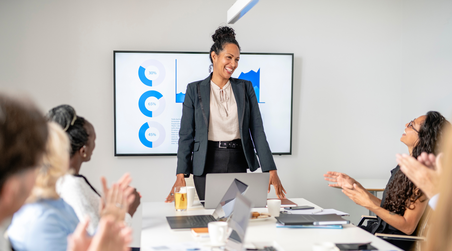 A business leader standing at a conference table during a meeting, smiling as colleagues applaud. A screen behind her shows performance charts, representing strategic decision-making, prioritization, and focused leadership.